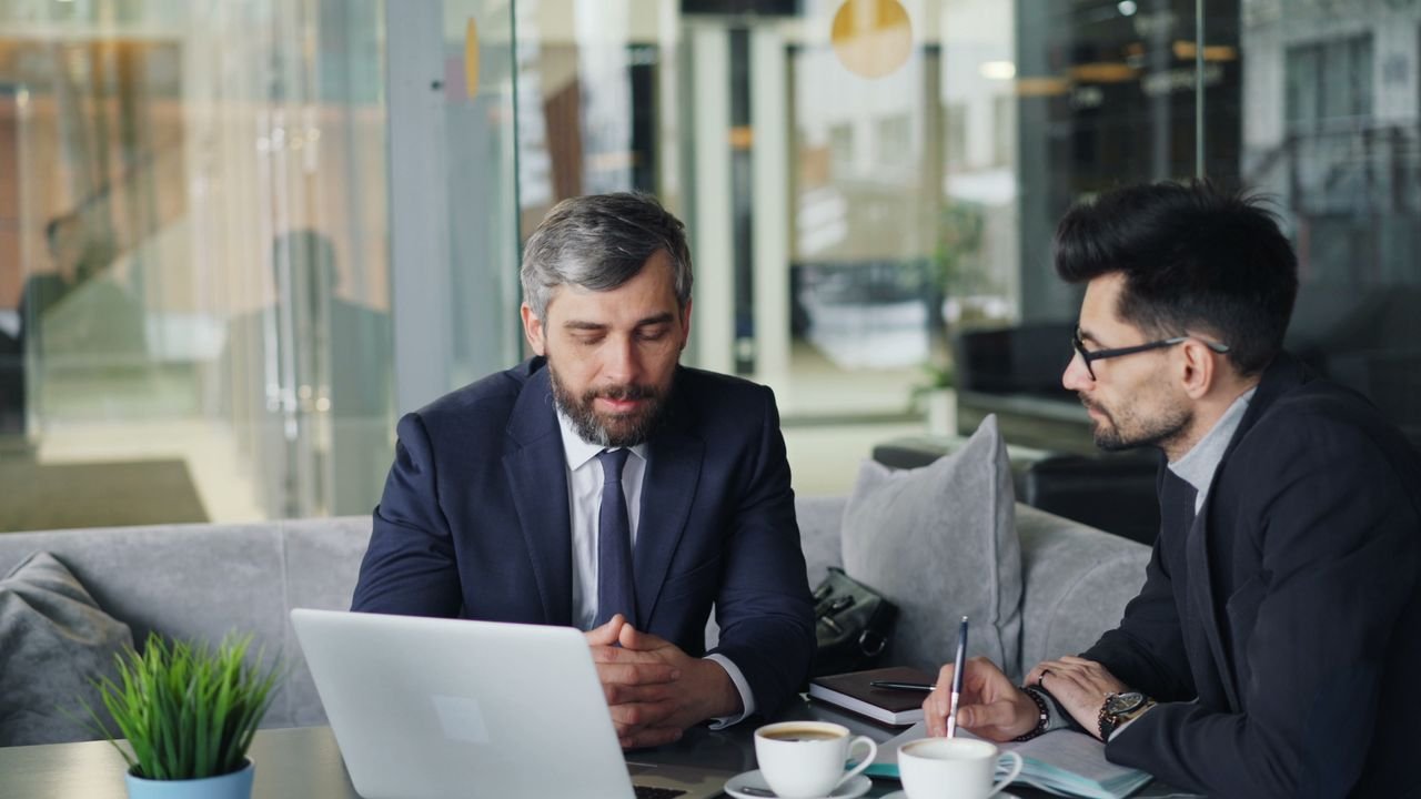 Contact Two businessmen in suits discussing work at a cafe with laptops and coffee.
